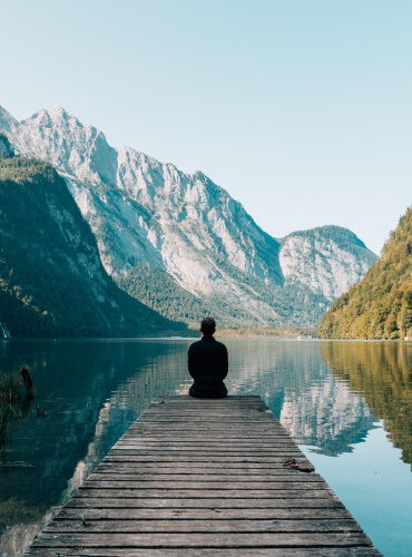 Eine Person sitzt allein auf einem Holzsteg mit Blick auf einen ruhigen See, umgeben von hohen Bergen und grünen Wäldern unter einem klaren Himmel.